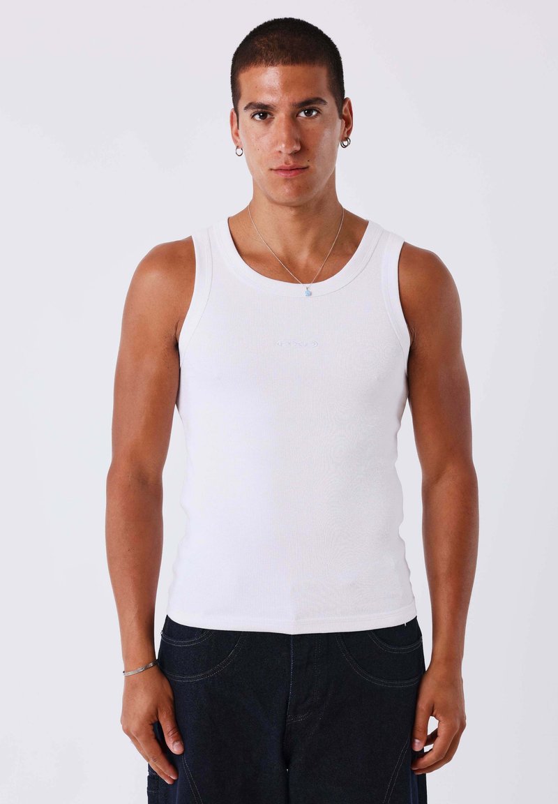 Jeune homme aux cheveux courts portant un haut blanc sans manches, un pantalon foncé, des boucles d'oreilles argentées en forme d'anneau et un collier, sur un fond uni.
