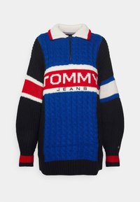 Pull en tricot bleu, noir et rouge avec un col blanc, une fermeture éclair devant et des motifs texturés. Présente un grand logo "TOMMY JEANS".