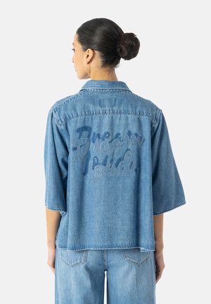 Kvinde med mørkt hår i en knold iført en løs lyseblå denimskjorte med teksten "Dream loud." broderet på ryggen og matchende denimbukser.