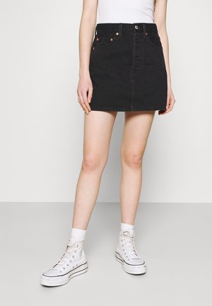 Minifalda de mezclilla negra con bolsillos frontales, botón de metal y detalles de costura. Llevada con zapatillas altas blancas y calcetines.