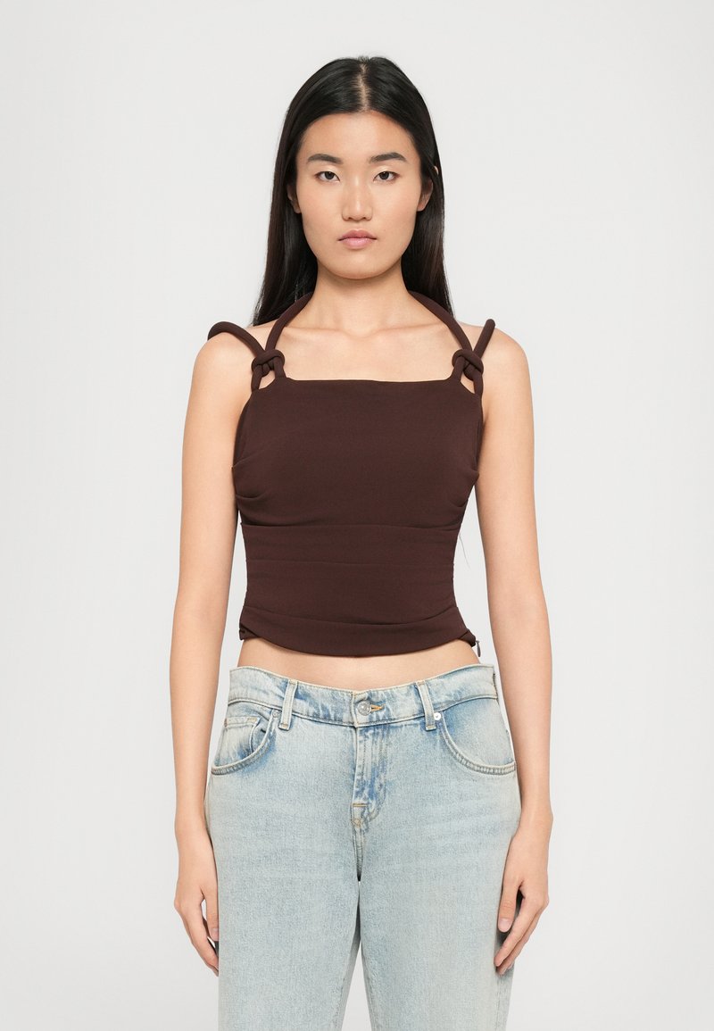 Bruine nauwsluitende tanktop met vierkante hals en geraffineerde zijkanten, voorzien van lusvormige schouderbanden. Gecombineerd met lichtblauwe hoge taille jeans.