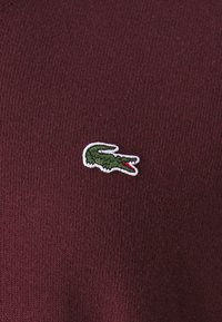 Burgundi színű pulóver, puha tapintással; a bal mellrészen egy fehér keretű zöld krokodil logóval.