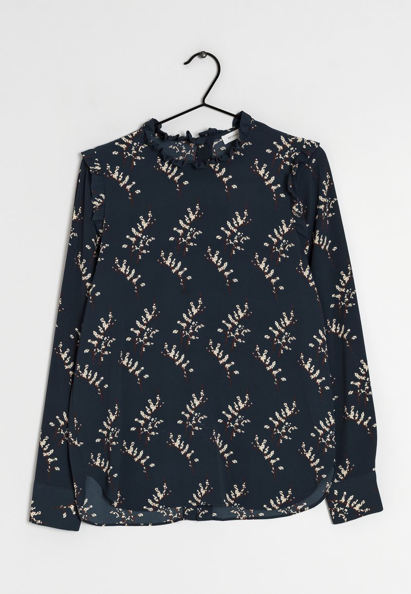 Blouse bleu marine à manches longues avec motif floral blanc et col volanté, suspendue sur un cintre noir sur fond blanc.