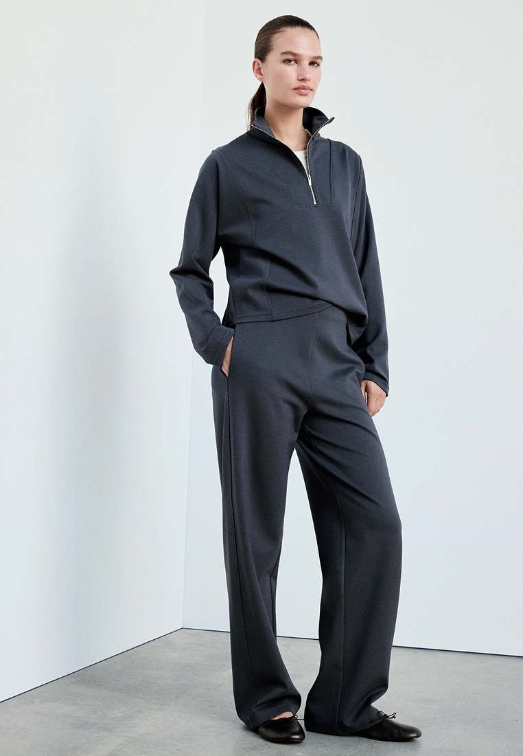 Femme debout, les mains dans les poches, portant un haut zippé gris foncé à coupe décontractée et un pantalon large assorti avec des chaussures plates noires.