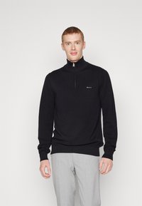 Zwarte gebreide trui met een hoge kraag en halve rits, met een textuurstof en een klein logo op de borst. Gedragen met lichtgrijze broeken.