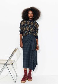 Pull en laine marron à motifs argyle avec des accents verts, associé à une jupe en plaid sombre. Des chaussettes bordeaux et des chaussures à semelle épaisse complètent le look.