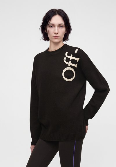 Personne aux cheveux courts et foncés portant un pull noir avec le mot "Off-" en grandes lettres crème sur l’épaule et un pantalon noir avec une fine bande bleue.