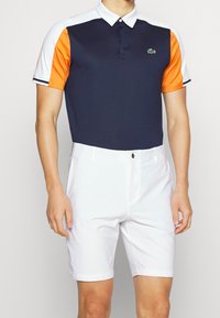 Marinblå polo skjorta med orange och vita ärmar, med krage och en grön logotyp. Kombineras med vita shorts som har en skräddarsydd passform.
