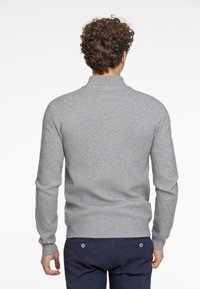 Maglione grigio a maglia con collo alto, maniche aderenti e orlo a coste. Texture morbida con dettagli minimi, indossato con pantaloni scuri.