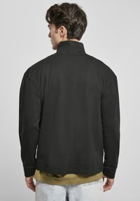 Zwarte lange mouwen shirt met een hoge kraag en een losse pasvorm, gemaakt van zachte stof. Gelaagd over een groene top met korte mouwen.