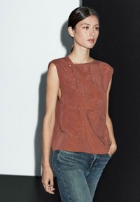 Rostfärgad ärmlös topp med en texturerad yta, som har en knappdetalj på ena sidan och en avslappnad passform, kombinerad med denimjeans.