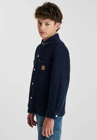 Tienerjongen met kort, krullend haar, gekleed in een marineblauw overhemd met knopen en een blauwe spijkerbroek, staande opzij tegen een effen achtergrond.