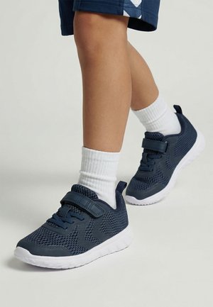 Kind trägt marineblaue Mesh-Sneaker mit weißen Sohlen, weiße Crew-Socken und marineblaue Shorts, steht vor hellem Hintergrund.
