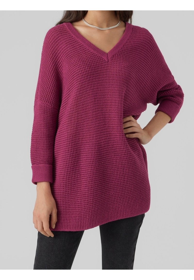 Femme portant un pull en maille violet oversize à col en V avec des manches retroussées et un pantalon noir, une main sur la hanche et l'autre détendue.