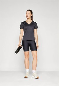 Juoda V formos iškirptės marškinėliai ir tamsūs šortai, derinami su baltais kojiniais ir smėlio spalvos sportiniais bateliais. Laiko juodą gertuvę. Lygios audinio tekstūros.