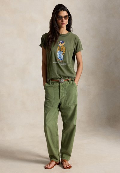 Γυναίκα με γυαλιά ηλίου, ελιάς πράσινο t-shirt με γραφικό αρκούδας, φαρδιά πράσινα παντελόνια με ζώνη και καφέ σανδάλια, στέκεται με τα χέρια στις τσέπες.