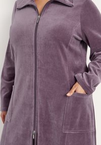 Lila fleece ytterplagg med dragkedja fram, långa ärmar och två framfickor; har en platt krage och mjuk textur.