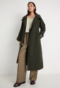 Casaco verde oliva, de botões duplos, com grandes lapelas e bolsos laterais. Tecido texturizado. Combinado com umas calças bege e sapatos escuros em bordô.