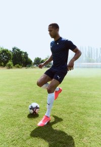 Athlète portant un maillot Puma bleu marine et un short noir, effectuant un coup de pied avec un ballon de football blanc sur l'herbe. Chaussures de football roses vives aux pieds.