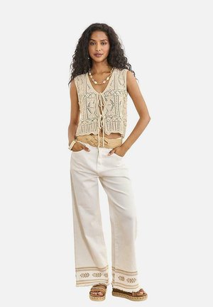 Mujer con chaleco beige de crochet con lazada al frente, pantalones blancos de pierna ancha con dobladillo bordado, cinturón tejido, sandalias y un collar de cuentas, de pie con las manos en los bolsillos.
