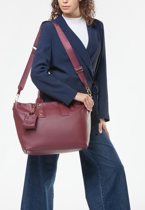 Sac à main en cuir bordeaux avec une texture lisse. Comprend un charme amovible, des poignées doubles et une bandoulière réglable. Design élégant et spacieux.