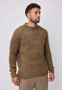 Homme aux cheveux courts bouclés et à la barbe, portant un pull texturé marron et un pantalon beige, debout devant un arrière-plan clair uni.