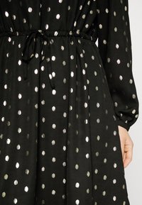 Vestido de tecido preto com bolinhas metálicas prateadas. Apresenta uma cintura franzida com cordão ajustável e mangas compridas. Textura suave.