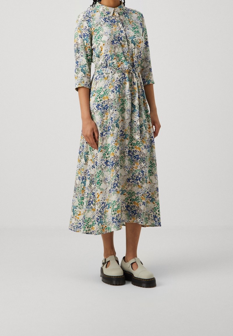 Femme portant une longue robe fleurie à manches trois-quarts avec un devant boutonné, associée à des chaussures beiges à plateforme épaisse.