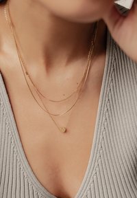 Drei geschichtete goldene Halsketten: eine dünne Kette, eine gedrehte Kette und eine kurze Kette mit einem kleinen Herzanhänger. Tragen auf einem grauen, gerippten Oberteil.
