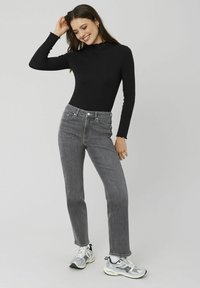 Body noir à manches longues avec un col montant, associé à un jean gris taille haute. Le modèle porte des baskets de sport argentées et se tient sur un fond neutre.