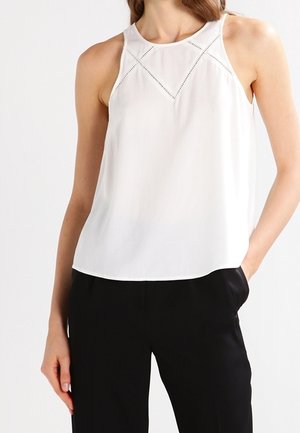 Hihaton valkoinen yläosa, jossa geometriset leikkauskuviot, yhdistetty mustiin housuihin, oikea käsi taskussa, yksiväristä taustaa vasten.