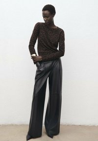 Top de manga larga marrón oscuro con estampado de leopardo, combinado con pantalones de cuero negros de pierna ancha. Los detalles destacados incluyen zapatos negros de punta y un reloj.