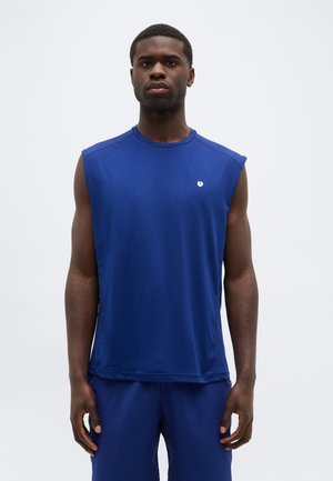 Άνδρας που φορά αμάνικη μπλε αθλητική μπλούζα και αντίστοιχο σορτς, στέκεται μπροστά σε απλό φωτεινό φόντο, κοιτάζοντας μπροστά με ουδέτερη έκφραση.