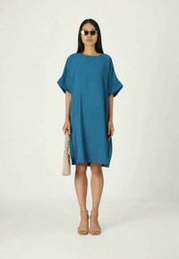 Ένα φορετό, κοντομάνικο φόρεμα σε τυρκουάζ ύφασμα, με στρογγυλή λαιμόκοψη. Φορεμένο με μπεζ σανδάλια και κρατώντας μια φωτεινή τσάντα.