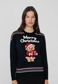 Marineblauer Pullover mit einem rot-weißen festlichen Strickmuster, das einen gestickten Bären in einem Weihnachtsmann-Outfit zeigt, der ein Geschenk hält und den Text "Frohe Weihnachten".