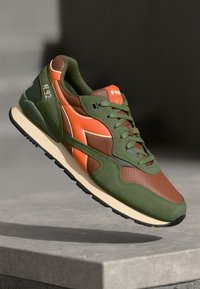 Green and brown sneaker with orange accents, featuring a mesh and leather upper, round toe, and textured detailing on the sides.