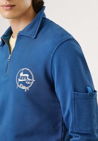 Mėlynas užtrauktukas džemperis, pagamintas iš tekstūruoto audinio, su emblemą turinčiu baltais užrašais ir kišene kairiajame rankovėje.