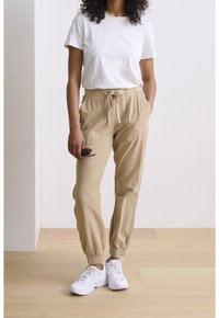 Beige Jogginghosen mit Gummibündchen und Seitentaschen, kombiniert mit einem schlichten weißen T-Shirt und weißen Sneakers. Minimalistischer, lässiger Stil.