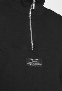 Svart tröja med hög krage, silverdragkedja och en rektangulär etikett på bröstet som visar varumärkets och platsinformation.