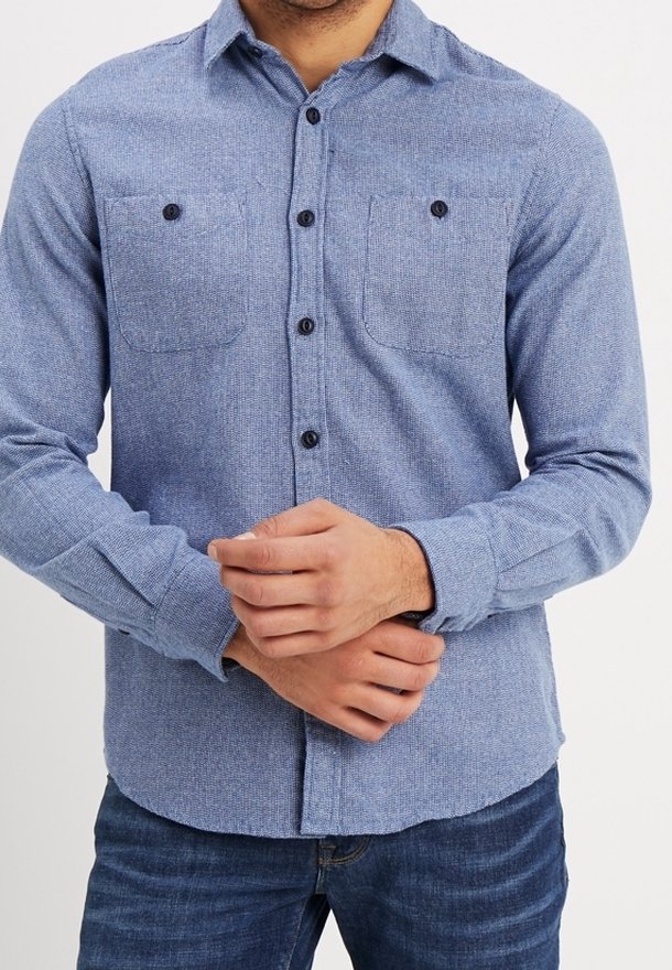 Camicia blu con bottoni realizzata in tessuto texturizzato, dotata di due tasche sul petto, bottoni neri e maniche lunghe con polsini abbottonati.