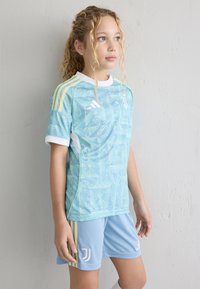 Lys blå ungdomsfotballdrakt har et virvelmønster på skjorten, med hvite og gule detaljer. Shortsen er ensfarget lys blå, med logodetaljer.