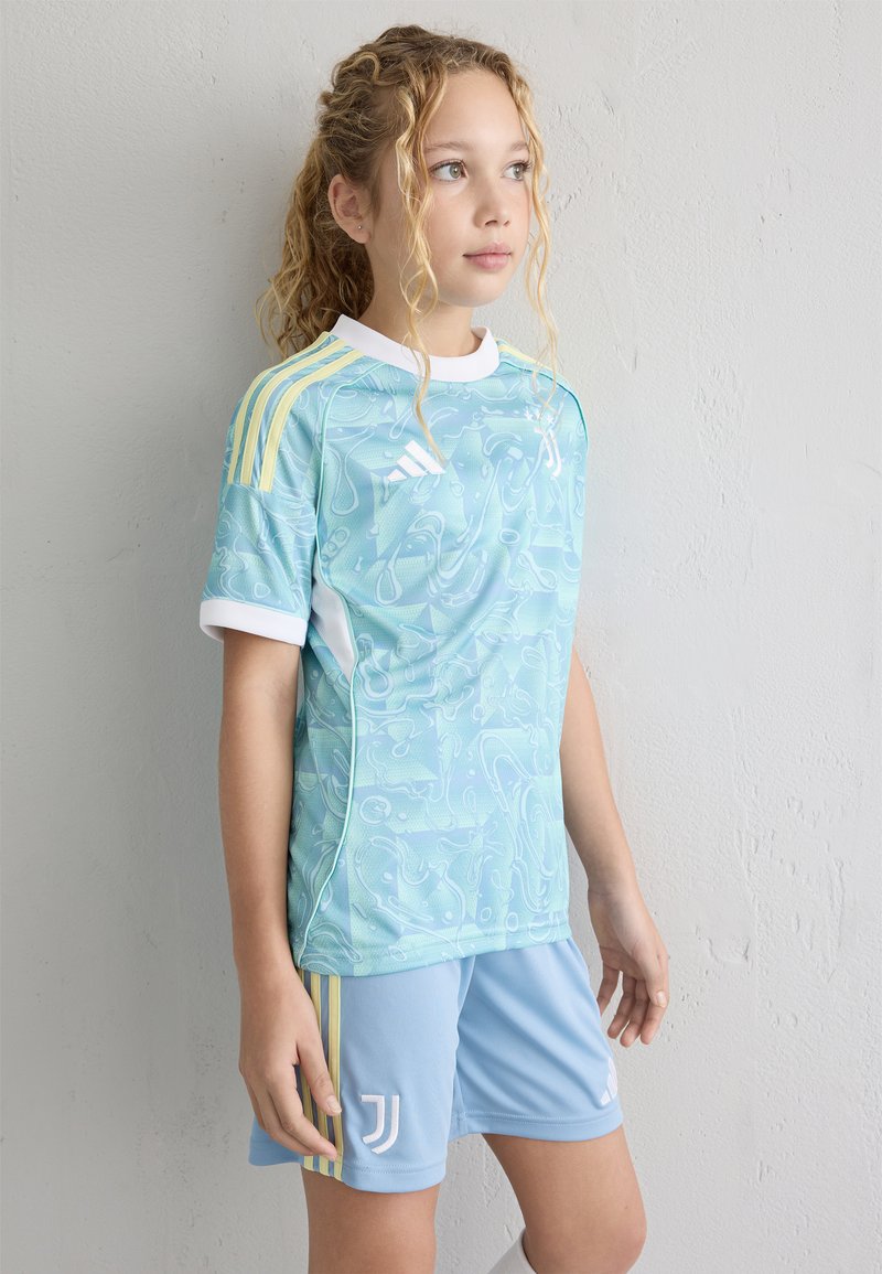 Lys blå ungdomsfotballdrakt har et virvelmønster på skjorten, med hvite og gule detaljer. Shortsen er ensfarget lys blå, med logodetaljer.