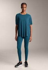 Teal, bő szabású póló, amelyhez illő leggings tartozik, puha textúrával és hosszabbított hátrésszel, fekete sportcipővel viselve.