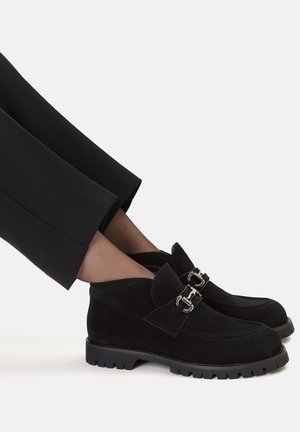 Czarne zamszowe mokasyny na grubych podeszwach ze srebrnymi klamrami, noszone z czarnymi spodniami i przezroczystymi brązowymi skarpetkami.