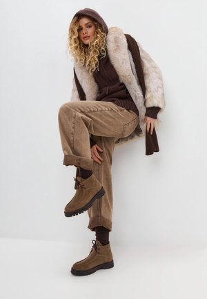 Braune Wildlederschuhen mit schwarzen Gummisohlen, kombiniert mit beigen Cordhosen und einem braunen Strickpullover unter einem Kunstpelzmantel.