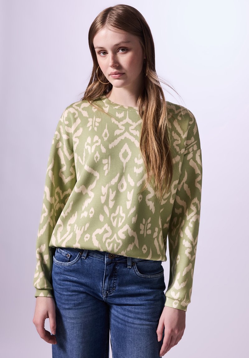 Vaaleanvihreä collegepaita, jossa on kermaisen värinen abstrakti kuvio, pyöreä kaula-aukko ja pitkät hihat, yhdistettynä sinisiin farkkuihin. Pehmeä kangas.