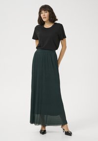 Maglietta nera a maniche corte abbinata a una lunga gonna plissettata di un verde scuro. L'outfit è completato da scarpe col tacco nere.