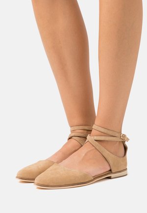 Pieds portant des ballerines pointues en suédine beige avec des sangles à la cheville et de petites boucles, présentés sur un fond blanc uni.