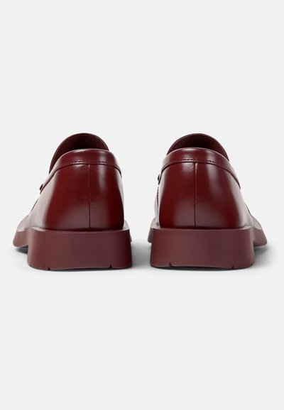 Par de mocasines de cuero pulido en rojo oscuro vistos desde atrás con suelas gruesas de goma sobre un fondo blanco liso.