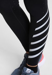 Leggings sportivi neri con un motivo laterale di strisce grigie a contrasto e piccole perforazioni, abbinati a scarpe da corsa grigie.